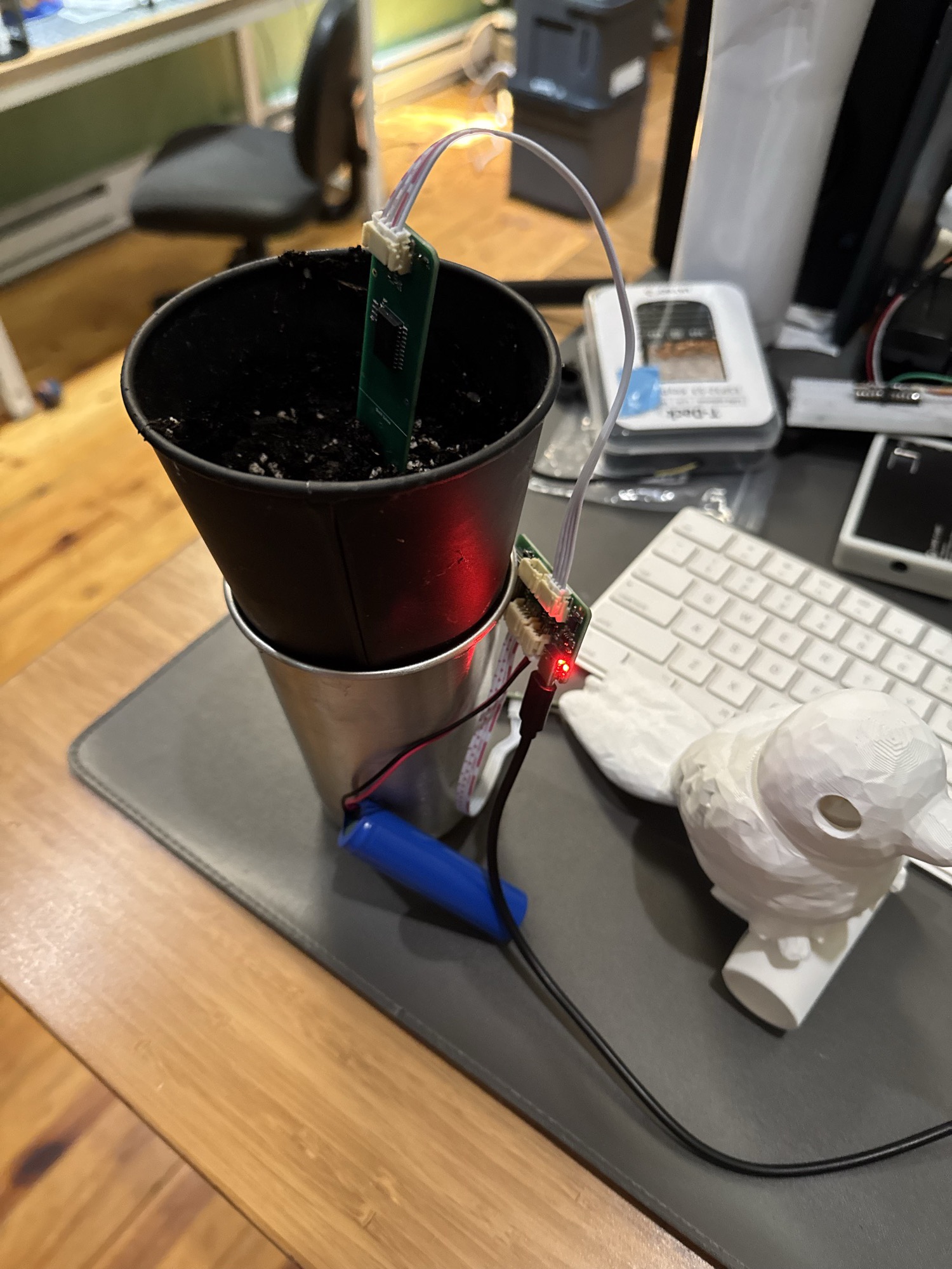 A green Prong sensor blade standing upright in a small pot of dark soil, connected by ribbon cable to a small board with a glowing red LED, powered by a blue lithium cell