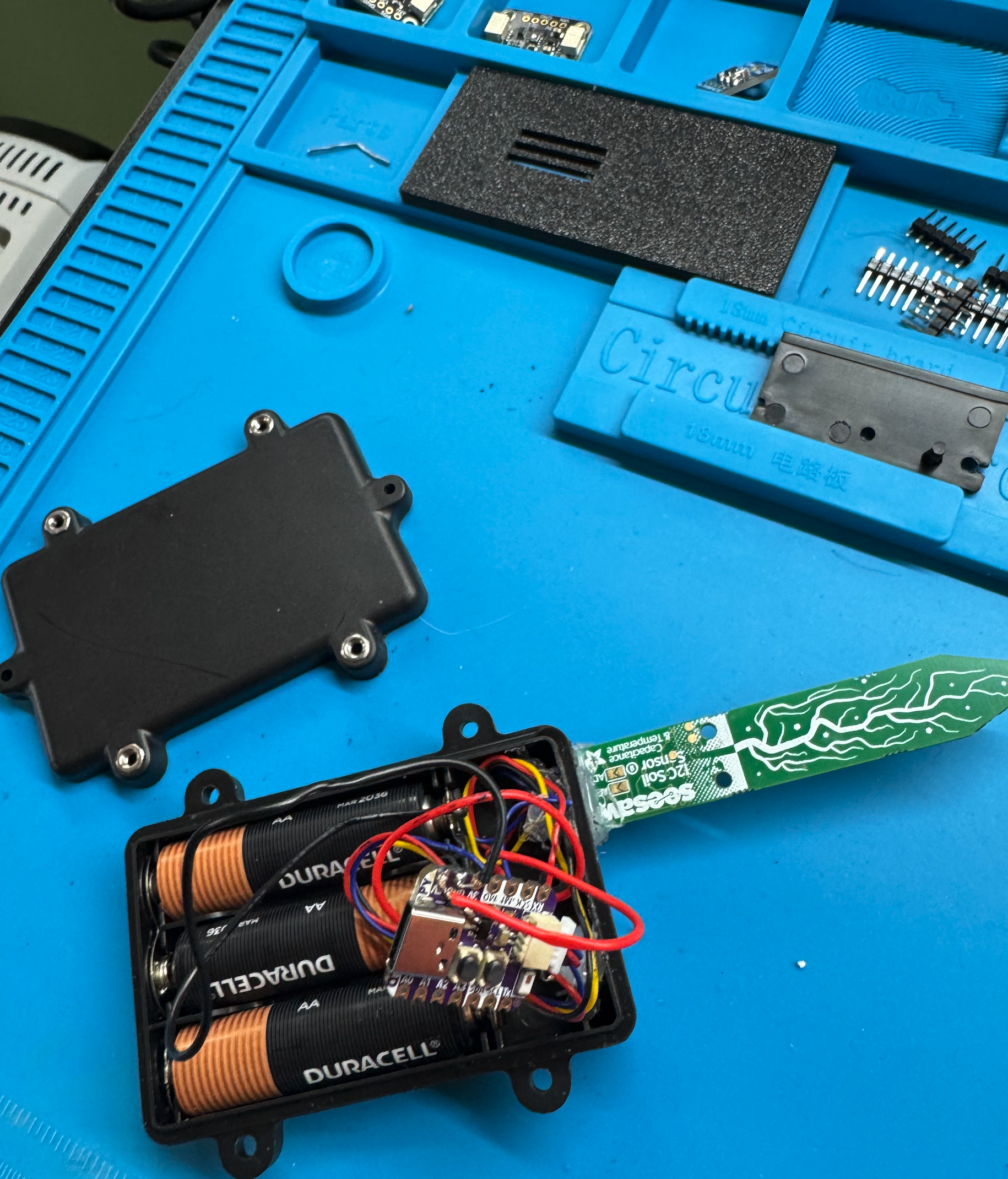 Aspirational Ani opened up on the soldering mat, QT Py crammed in with batteries, soil sensor sticking out the side